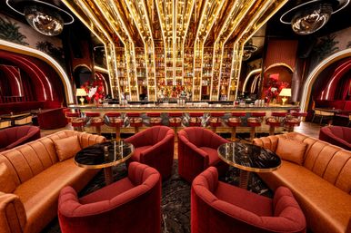 Glamorous gold-lit cocktail lounge interior with mirrored, bottle-lined back bar, red velvet and leather seating, marble tables and brass barstools.