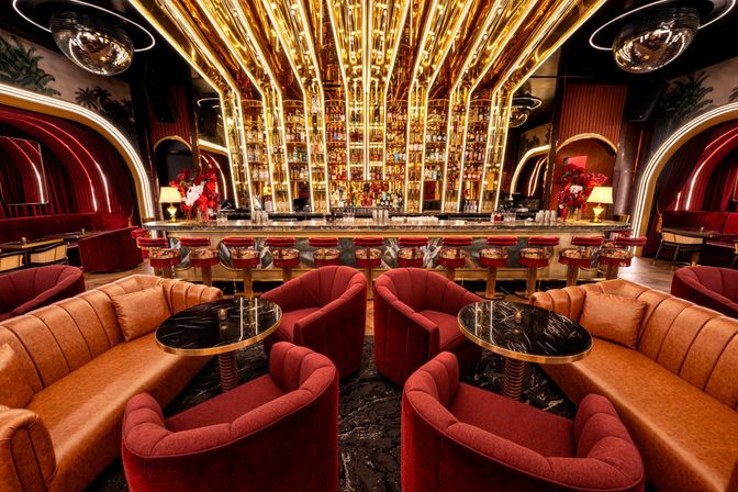 Luxurious Art Deco cocktail lounge interior with gold-lit mirrored back bar, red velvet chairs, tan leather sofas, black marble tables and disco balls
