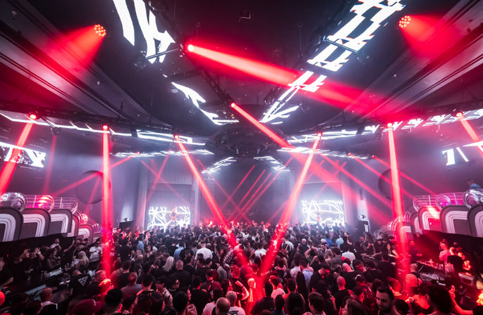 Packed nightclub interior with a dancing crowd pulsing under red laser beams and geometric LED visuals over the stage, energetic late-night party scene