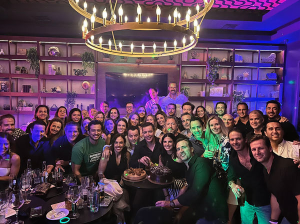 Cheerful large group celebrating in an indoor lounge or bar, gathered around a table with two birthday cakes, wine glasses and bottles, lit by a round chandelier and purple-blue party lighting with decorative shelving in the background.