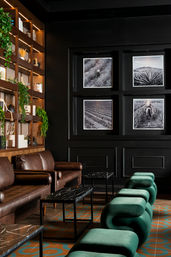 Moody modern boutique hotel lounge with brown leather sofas, emerald-green sculptural velvet stools, black marble tables, wood shelving with plants, and four black-and-white framed landscape photos on a dark wall.
