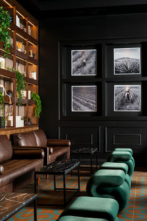 Moody modern boutique hotel lounge with brown leather sofas, emerald-green sculptural velvet stools, black marble tables, wood shelving with plants, and four black-and-white framed landscape photos on a dark wall.