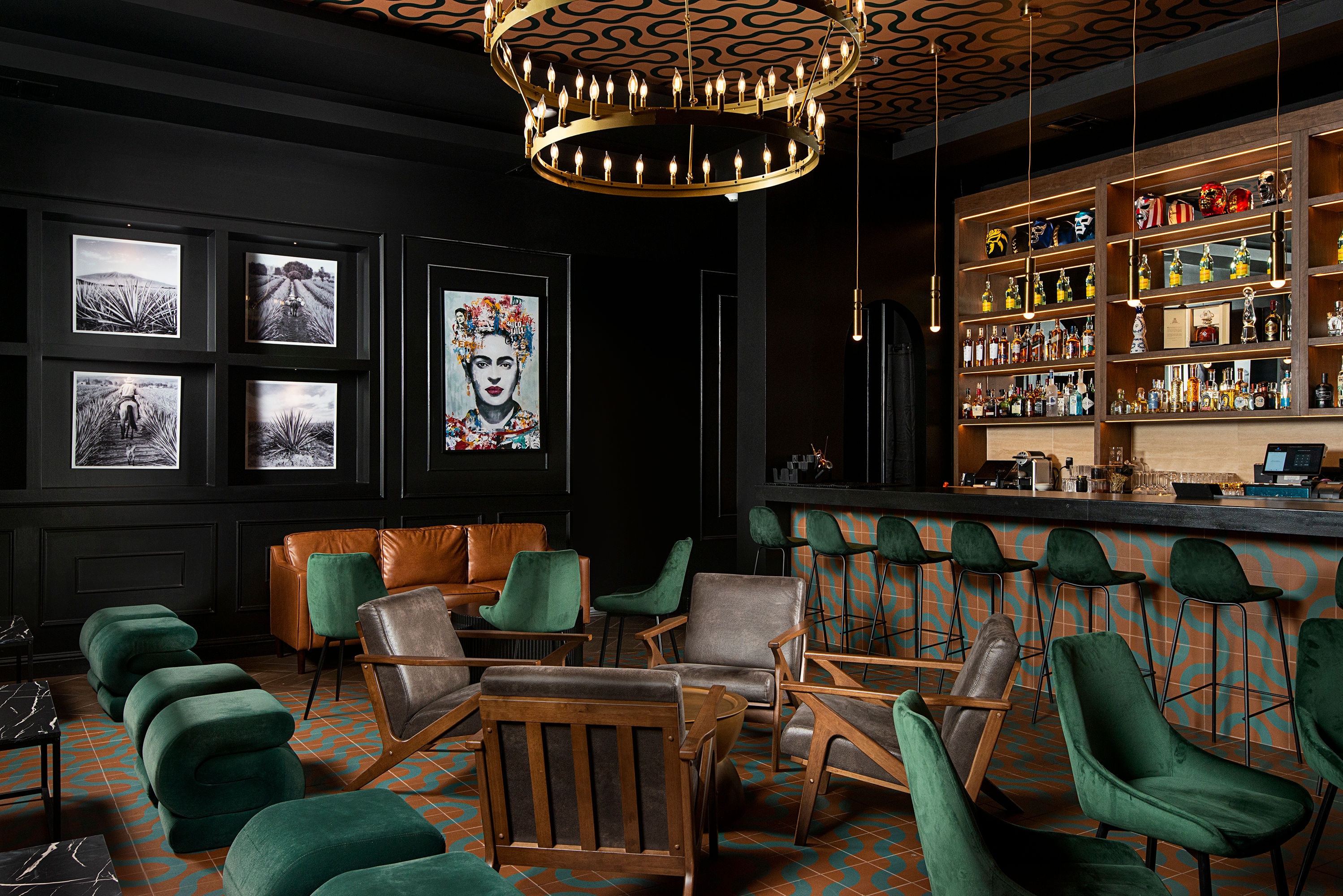 Boutique hotel cocktail lounge with emerald velvet seating, cognac leather sofa, mid-century wood armchairs, patterned tile floor, illuminated backbar and brass ring chandelier, colorful portrait art on dark paneled wall.