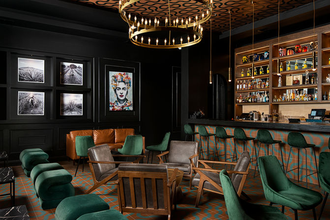 Boutique hotel cocktail lounge with emerald velvet seating, cognac leather sofa, mid-century wood armchairs, patterned tile floor, illuminated backbar and brass ring chandelier, colorful portrait art on dark paneled wall.