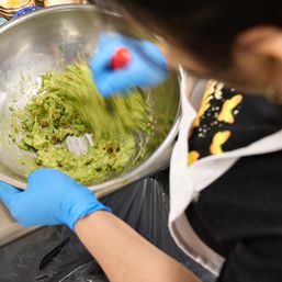Blue-gloved hands vigorously mixing chunky guacamole in a stainless-steel bowl during kitchen food prep.