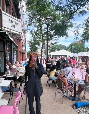 Person in a jumpsuit and beret holding a pink drink in front of a busy sidewalk café and summer street festival with outdoor dining, market tents, and city trees.