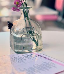 Tequila bottle repurposed as a vase holding pink daisy-like blooms on a white café table next to a laminated menu — bright, casual tabletop centerpiece.