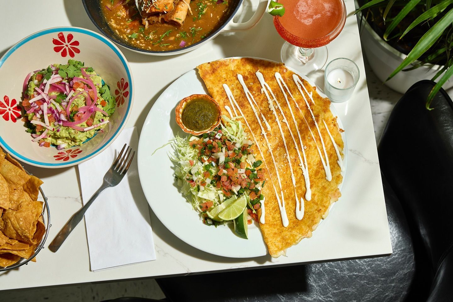 Top-down view of a crispy cheese quesadilla drizzled with crema, served with shredded cabbage pico de gallo, lime wedges and green salsa, plus a bowl of guacamole, tortilla chips and a pink margarita on a white table — Mexican-style meal.