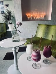 Cozy cafe corner with white subway-tile wall and glowing candles, green textured chairs, a single pink flower in a glass bottle, and three colorful frozen cocktails on small white tables.