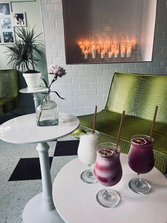 Cozy cafe corner with white subway-tile wall and glowing candles, green textured chairs, a single pink flower in a glass bottle, and three colorful frozen cocktails on small white tables.