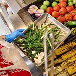 Gloved cook holding a tray of charred scallions and green onions over a sizzling grill of roasted tomatoes, halved red onions, tomatillos and charred corn on the cob.
