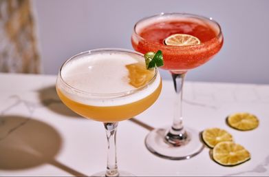 Two vibrant cocktails in coupe glasses on a white marble countertop — a frothy yellow drink with a lime twist and a red frozen cocktail garnished with a dried lime slice, lime rounds nearby.