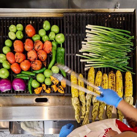 Top-down view of a commercial grill piled with charred tomatoes, green tomatillos and chiles, purple onions, small orange peppers, long green onions, and braided-husk corn on the cob being flipped with tongs by gloved hands.