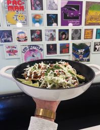 Hand holding a white enamel pan of loaded tacos topped with shredded cabbage, crema, pico de gallo, avocado slices and lime wedges, set against a colorful gallery wall of framed album-style art.