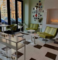 Bright urban lounge with green retro-chic chairs, white round tables, black-and-white checker floor, potted plants and large windows overlooking a city street