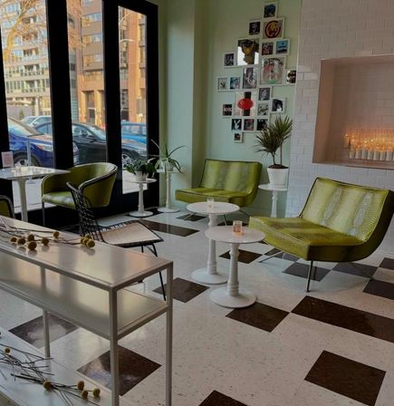 Bright urban lounge with green retro-chic chairs, white round tables, black-and-white checker floor, potted plants and large windows overlooking a city street
