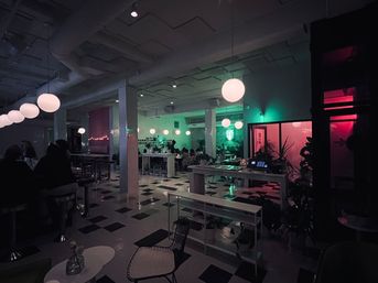 Neon-lit modern cafe interior with round pendant lights, black-and-white checkered floor, communal tables and diners bathed in green and pink ambient lighting