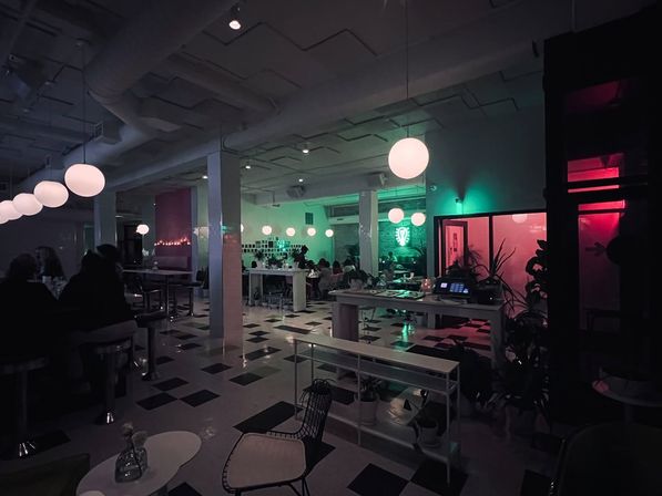 Neon-lit modern cafe interior with round pendant lights, black-and-white checkered floor, communal tables and diners bathed in green and pink ambient lighting