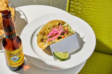 Crispy fried taco in a metal holder topped with pickled red onions, salsa and shredded cabbage, served with a lime wedge on a white plate, a beer bottle and basket of tortilla chips on a sunny outdoor table.