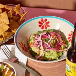Colorful Mexican-style guacamole topped with pickled red onions, cilantro and diced tomatoes in a floral bowl, served with crispy tortilla chips and a bottled beer on a wooden table
