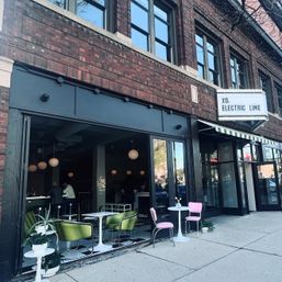 Downtown brick storefront café with open-air seating — green lounge chairs, pink bistro seats, white round tables, globe pendant lights and a striped awning on a city sidewalk.