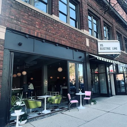 Downtown brick storefront café with open-air seating — green lounge chairs, pink bistro seats, white round tables, globe pendant lights and a striped awning on a city sidewalk.