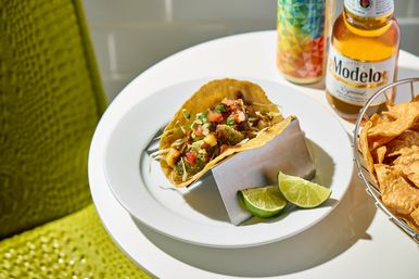 Crispy corn-shell taco topped with pico de gallo and shredded slaw on a white plate with lime wedges, a basket of tortilla chips and a cold beer on a sunlit table.