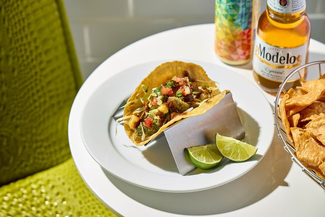 Crispy corn-shell taco topped with pico de gallo and shredded slaw on a white plate with lime wedges, a basket of tortilla chips and a cold beer on a sunlit table.