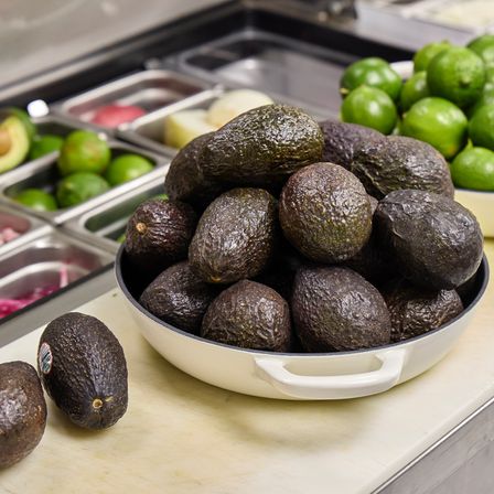 Pile of ripe Hass avocados in a white bowl on a restaurant prep counter, ready for guacamole, with bright limes and stainless-steel ingredient bins in the background