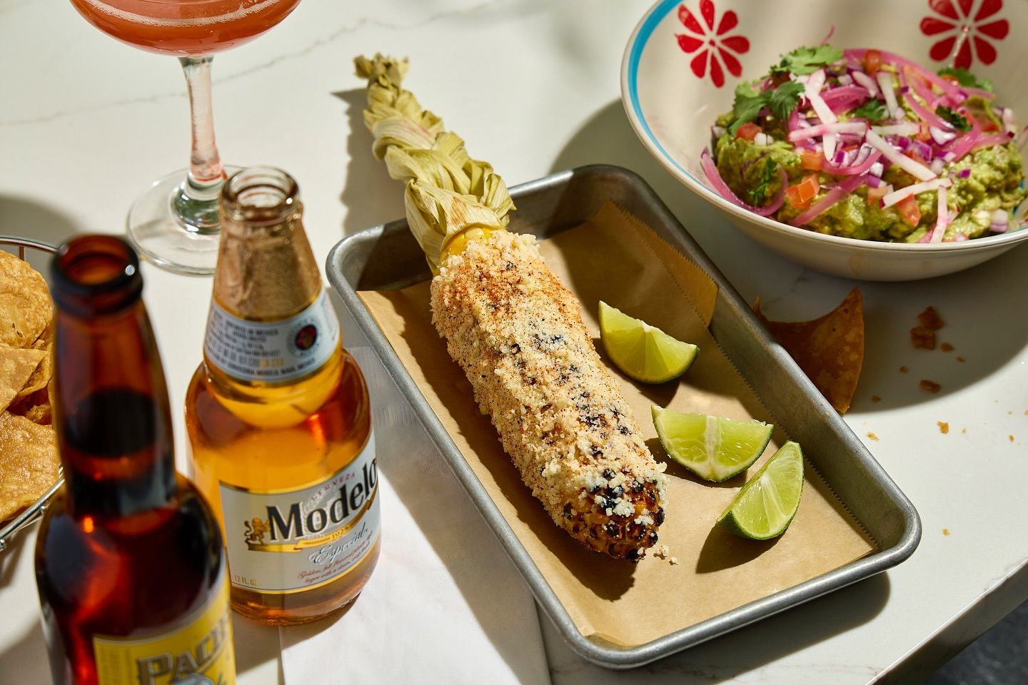 Taqueria-style elote (Mexican street corn) grilled and coated in crumbly cheese and spices on a metal tray with lime wedges, alongside a bowl of guacamole with pickled onions, tortilla chips and bottled beer on a white marble table.