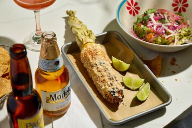 Taqueria-style elote (Mexican street corn) grilled and coated in crumbly cheese and spices on a metal tray with lime wedges, alongside a bowl of guacamole with pickled onions, tortilla chips and bottled beer on a white marble table.