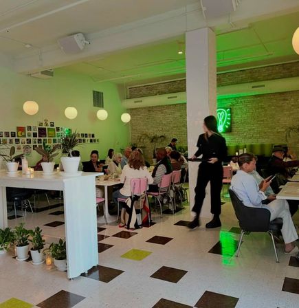 Busy modern urban café interior with diners at communal tables, pink chairs, globe pendant lights, potted plants, brown-and-lime tiled floor, green neon heart sign and a server carrying a tray.