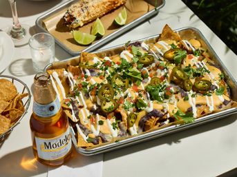 Crowd-pleasing tray of Mexican-style loaded nachos topped with queso, refried beans, sour cream, pico de gallo, sliced jalapeños and cilantro, served with grilled elote and a beer on the table.