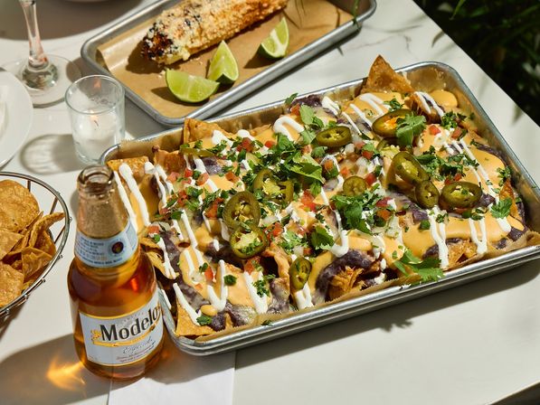 Crowd-pleasing tray of Mexican-style loaded nachos topped with queso, refried beans, sour cream, pico de gallo, sliced jalapeños and cilantro, served with grilled elote and a beer on the table.