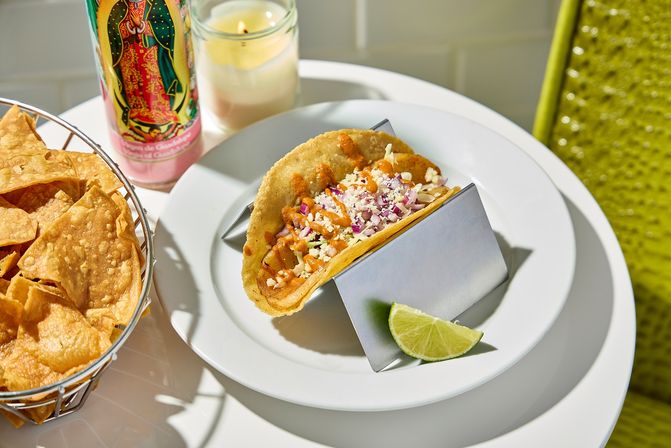 Crispy Mexican street taco with shredded cheese, diced onion and spicy sauce on a white plate with lime wedge, tortilla chips basket and a colorful devotional candle nearby