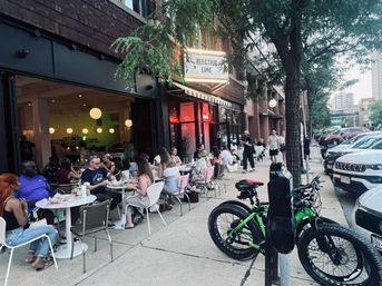 Bustling downtown sidewalk cafe patio at dusk, groups dining at outdoor tables under round pendant lights, neon sign over the entrance, tree-lined sidewalk with parked cars and green e-bikes locked to a meter.