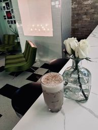 Iced latte topped with foam and cinnamon on a white marble countertop beside a glass vase of white roses in a cozy cafe interior with green vintage chairs and black-and-white checkered floor.