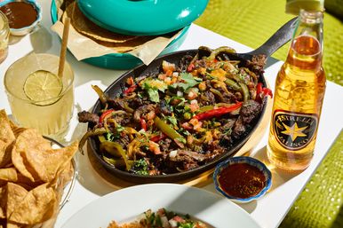 Sizzling Mexican steak fajitas in a cast-iron skillet with sautéed bell peppers and pico de gallo, served with tortilla chips, a lime margarita, and a bottle of Mexican beer on a bright outdoor table