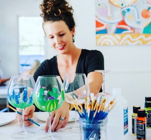 Smiling person painting colorful cactus and floral designs on wine glasses in a bright art studio, with jars of brushes and paint bottles on the table — hand-painted glassware workshop
