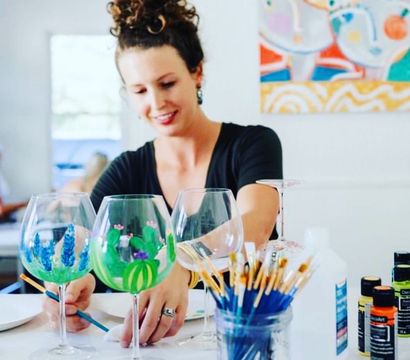 Smiling person painting colorful cactus and floral designs on wine glasses in a bright art studio, with jars of brushes and paint bottles on the table — hand-painted glassware workshop