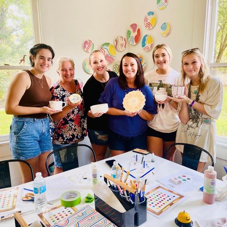 Six women in a bright craft studio holding hand-painted pottery and cups during a fun ceramic painting workshop, with brushes, paints and colorful wall plates on display.