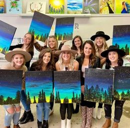 Smiling women in an indoor painting studio holding matching acrylic canvases of a blue-green sunset over silhouetted pine trees — group paint night.