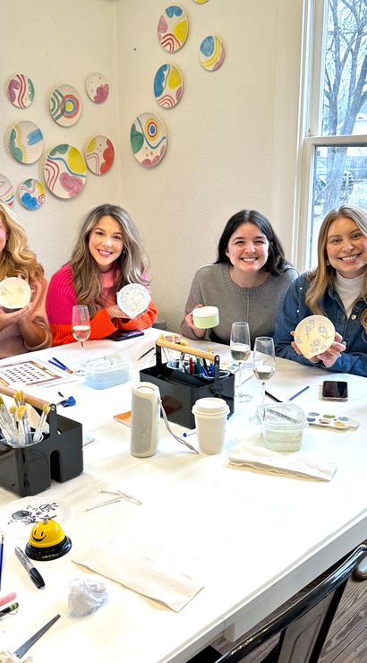Cheerful group of six women at a pottery-painting workshop in a bright art studio, seated at a white table holding painted ceramic bowls and cups with brushes, paints and colorful wall plates visible.