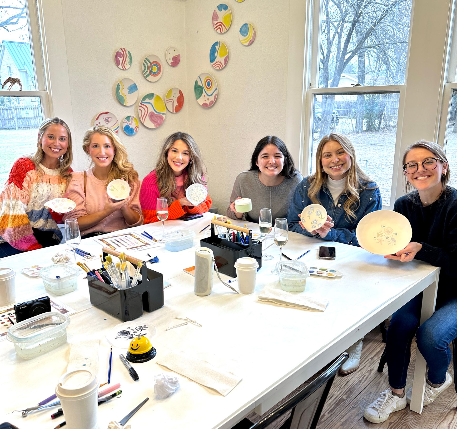 Cheerful group of six women at a pottery-painting workshop in a bright art studio, seated at a white table holding painted ceramic bowls and cups with brushes, paints and colorful wall plates visible.