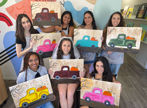 Seven women at an indoor art studio painting party hold colorful canvases of vintage pickup trucks carrying pumpkins; central guest wears a veil, bright fall-themed artwork and smiles.