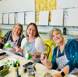 Three smiling women in paint-splattered aprons painting small craft ornaments at a bright art studio workshop, holding brushes and palettes with blank canvases and paint-splattered shelves in the background.