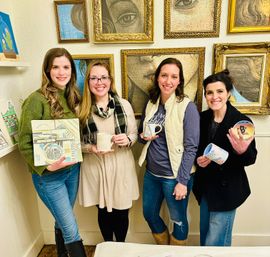 Four smiling women in casual winter outfits holding hand-painted ceramic mugs and a decorative tile inside a bright pottery painting studio with framed artwork on the wall.