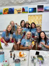 Nine women in matching teal shirts pose smiling in an art studio paint-and-sip session, holding hand-painted wine glasses amid bright canvas paintings and painting supplies on the table.