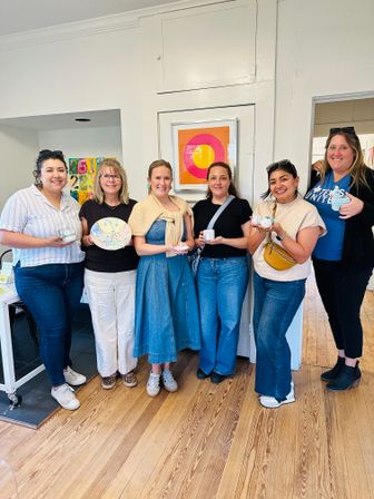 Six smiling women in casual denim standing in a bright art studio with wooden floors and colorful wall art, each holding hand-painted ceramics from a pottery painting workshop.