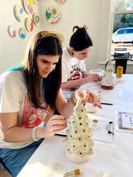 Two young women painting ceramic Christmas trees at a sunlit craft studio table by a window, brushes and paint palettes scattered around; one wears a glitter "Sweet Sixteen" headband and has blue hair streaks.